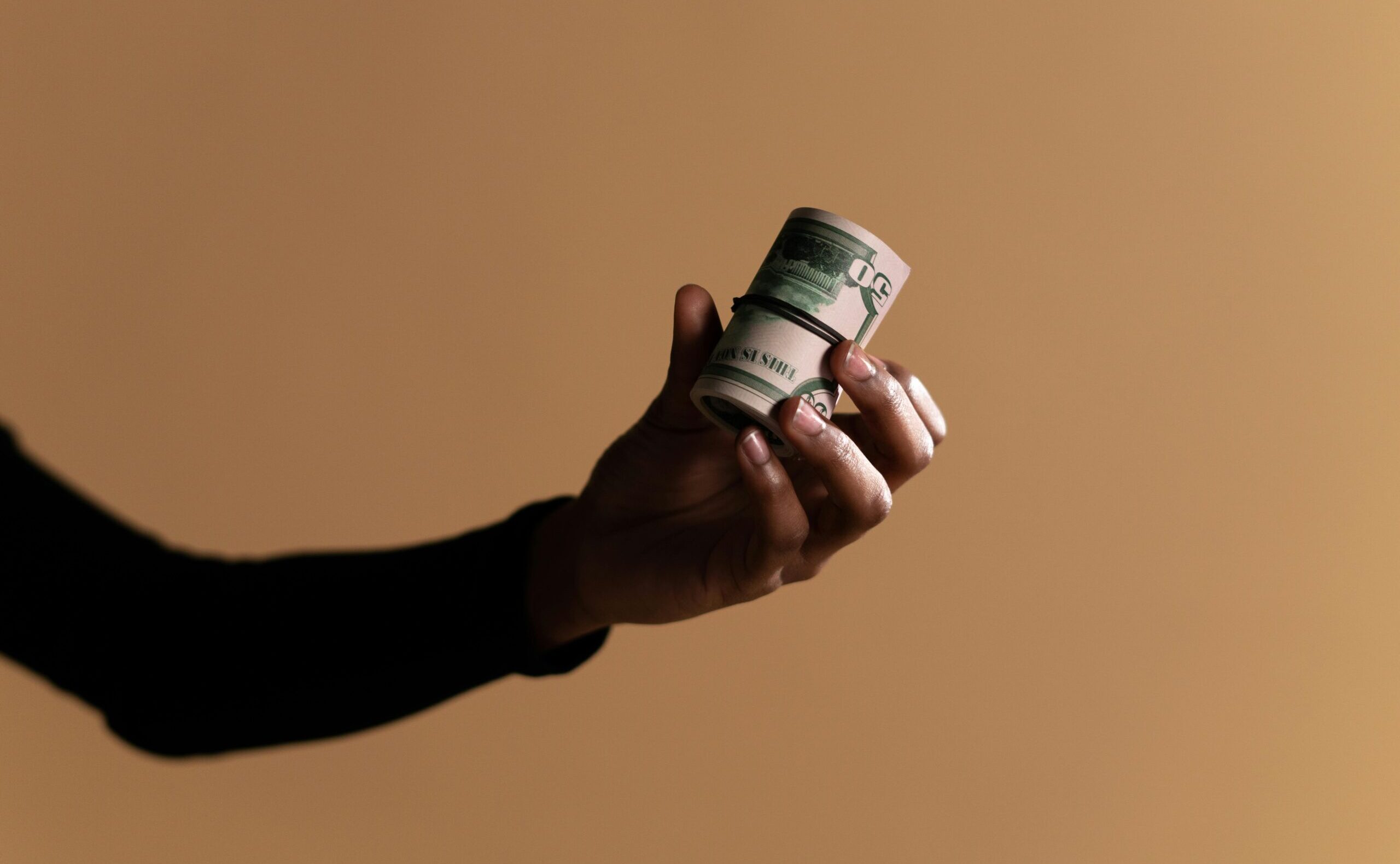 A close-up image showing a hand holding a roll of money against a beige background, ideal for finance themes.